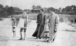 Two women and 3 boys in early 20th century clothing with a kite on the beach, a log cabin in the distance